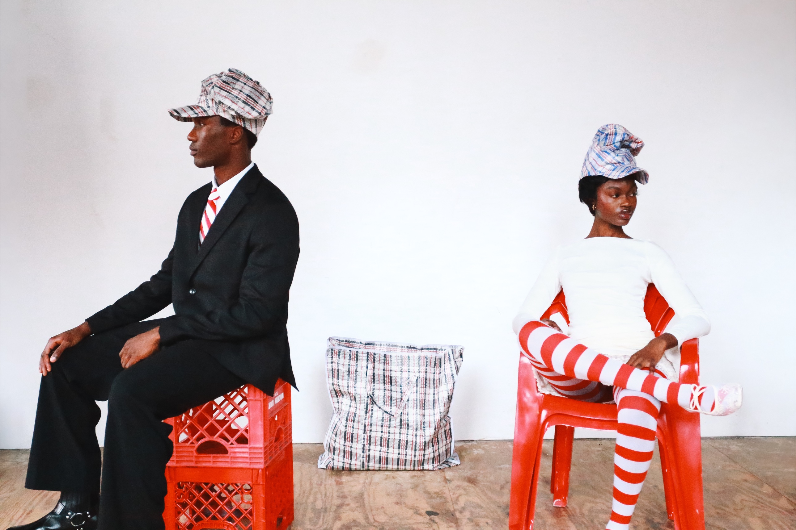 Two models wearing Homecoming Hats seated on a red milk crate and a red chair, a Ghana Must Go bag between them